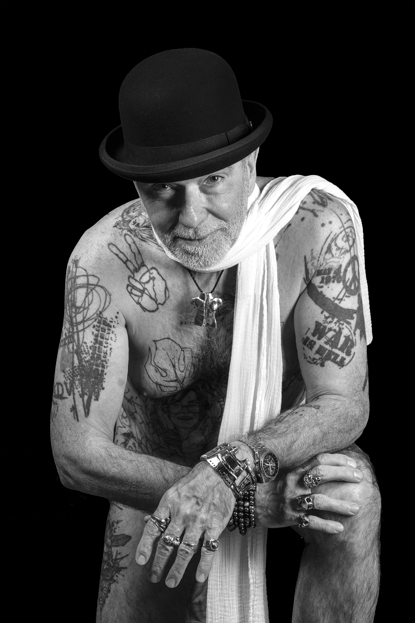 Le corps comme langage Portrait artistique en noir et blanc d’un homme tatoué, le visage visible sous un chapeau noir, avec une longue écharpe blanche couvrant le bas du corps.