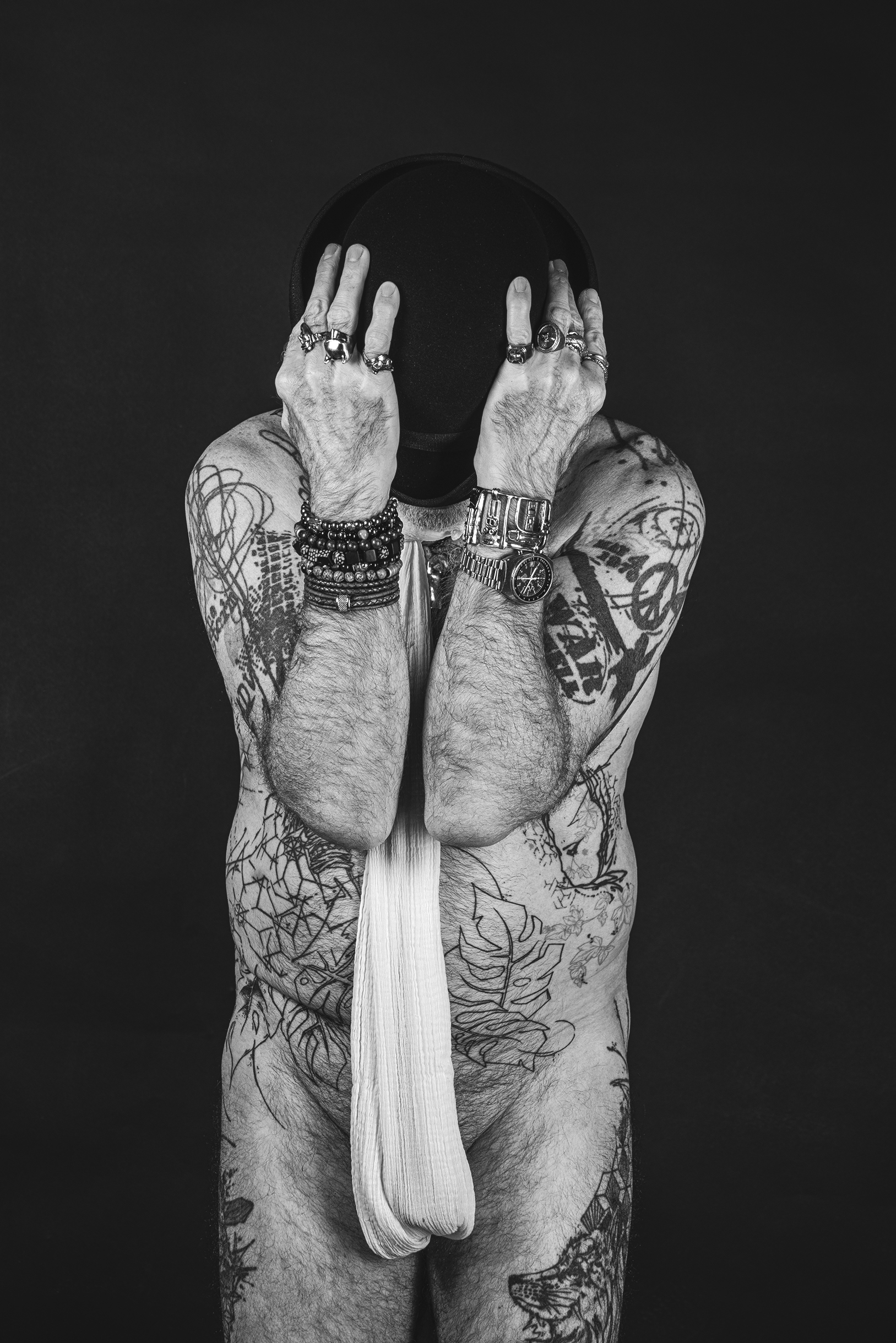 Le visage absent Homme tatoué en noir et blanc, visage caché par un chapeau et ses mains, portant des bijoux et un long tissu blanc sur fond noir.